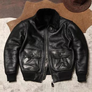 Può includere: Giacca bomber in pelle nera con collo in pelliccia nera, due tasche frontali e chiusura con cerniera. La giacca ha orlo e polsini a coste. Un cappello da cowboy marrone è visibile sullo sfondo.