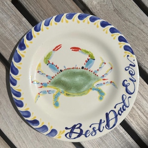 May include: A round, white ceramic plate with a blue crab illustration. The crab is green, blue, and red. The plate has a blue and yellow decorative border and the words "Best Dad Ever" in blue script.