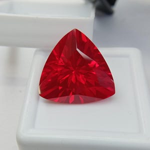 Op de afbeelding: Een levendige rode edelsteen geslepen in een driehoekige vorm met meerdere facetten die het licht reflecteren. De steen is geplaatst op een witte vierkante displaybasis, wat de dieprode kleur en scherpe, geometrische randen benadrukt.