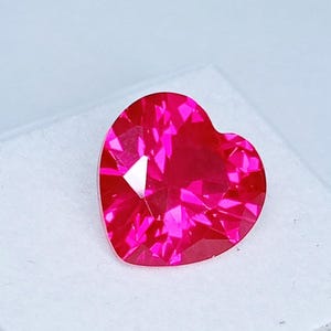 May include: A heart-shaped gemstone with a vibrant, saturated hot pink color. The stone features a faceted cut that creates multiple reflective surfaces, resulting in a brilliant, sparkling appearance with sharp geometric light patterns throughout.