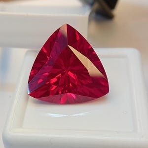 May include: A vibrant, deep red gemstone cut into a triangular shape with multiple facets that reflect light. The stone is positioned on a white square display base, showcasing its translucent quality and sharp, geometric edges.