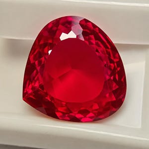 May include: A vibrant, deep red gemstone cut into a triangular shape with multiple facets. The stone exhibits a high level of clarity and brilliance, reflecting light across its surface. It is positioned against a plain, light-coloured background.