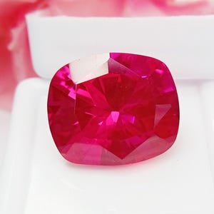 May include: A vibrant, cushion-cut gemstone with a deep, saturated red color and multiple triangular facets that create a brilliant, reflective surface. The stone is positioned centrally, showcasing its symmetrical shape and intense, vivid red hue.