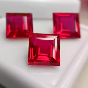 Può includere: Tre pietre preziose rosso vivo a taglio quadrato con superfici sfaccettate. Ogni pietra presenta una forma geometrica netta con bordi chiari e riflettenti e un colore cremisi saturo, disposte in una formazione triangolare.
