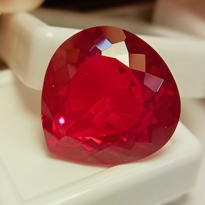 Peut inclure: Une pierre précieuse à facettes en forme de cœur d'une couleur rouge profond et vibrante. La pierre présente de nombreuses facettes triangulaires et en losange qui reflètent la lumière, créant un aspect brillant et étincelant sur sa surface polie.