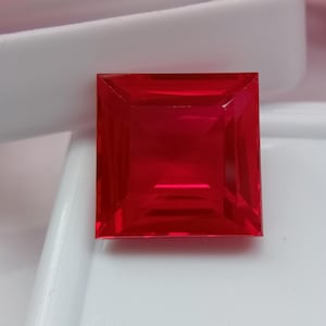 Può includere: Una pietra preziosa a taglio quadrato di un colore rosso vibrante e traslucido. La pietra presenta una superficie sfaccettata che crea motivi geometrici e riflessi di luce sulla parte superiore. La pietra preziosa è posizionata su una superficie bianca liscia.