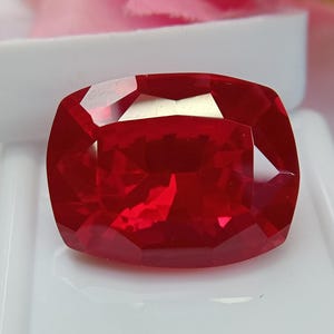 Könnte beinhalten: Ein leuchtend tiefroter Edelstein im Kissenschliff mit zahlreichen Facetten, die das Licht auf seiner Oberfläche reflektieren. Der Stein zeigt einen satten, kräftigen Karmesinton und ist vor einem glatten, weißen Hintergrund platziert.