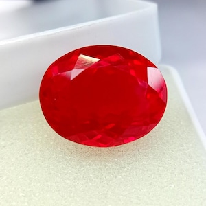 Könnte beinhalten: Ein leuchtender, oval geschliffener Edelstein mit tiefroter Farbe und facettierter Oberfläche. Der Stein hat ein poliertes Finish, das das Licht über die gesamte Oberfläche reflektiert und einen brillanten, gesättigten roten Farbton zeigt.