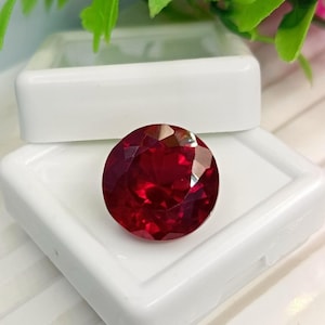 May include: A round, faceted, deep red gemstone with a brilliant cut, displaying multiple triangular and kite-shaped facets that reflect light. The gemstone is positioned on the open lid of a small, square, white plastic display box.
