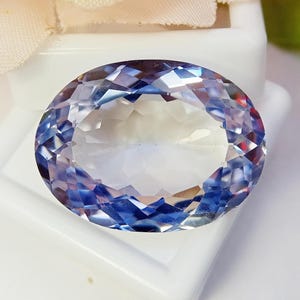 May include: An oval-cut gemstone featuring a faceted surface that displays a gradient of translucent white, soft violet, and deep blue hues. The stone is positioned on a white display base, showcasing its reflective clarity and geometric cut.