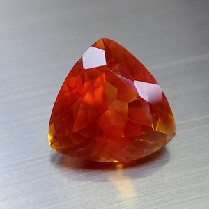 Pode incluir: Uma pedra preciosa de corte triangular com uma paleta de cores laranja vibrante e vermelho profundo. A pedra apresenta múltiplas facetas geométricas que refletem a luz, criando uma aparência brilhante com tons de âmbar quente e carmesim.