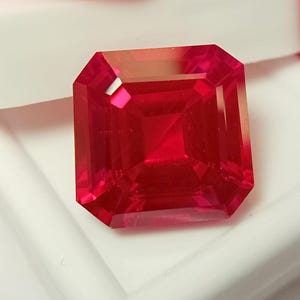 Puede incluir: Una piedra preciosa de corte cuadrado con un color rojo vibrante y translúcido, con superficies facetadas que crean reflejos geométricos nítidos. La piedra presenta un acabado pulido y claro con bordes definidos y una simetría de refracción.
