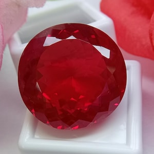 May include: A round, faceted, deep red gemstone with a brilliant cut, displaying multiple triangular and diamond-shaped facets that reflect light across its surface. The stone is positioned centrally, resting on a small, square white display base.