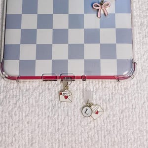 Puede incluir: Una funda rectangular para tableta con un patrón de cuadros azul claro y blanco. Un dije de cadena de metal dorado cuelga de la parte superior, adornado con un corazón azul, un lazo rosa, una perla y un dije circular con la letra S. Dos dijes de sobre con corazones rojos cuelgan del borde inferior, uno con la letra L.