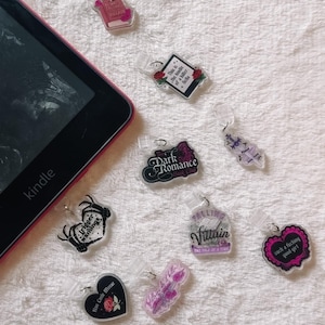 Puede incluir: Una colección de dijes de libros de acrílico con varios diseños y texto, incluyendo "Dark Romance", "Trigger Warnings" y "Villain". Los dijes son de varias formas y colores, con un lector de libros electrónicos Kindle en el fondo.