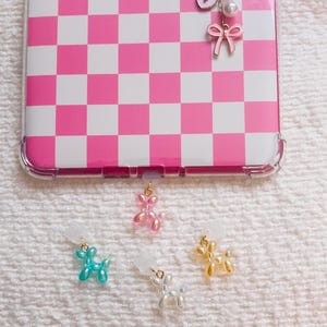 Può includere: Una cover per telefono con motivo a scacchi rosa e bianco dotata di un ciondolo dorato con fiocco, cuore e la lettera S. Quattro piccoli ciondoli a forma di cane palloncino in rosa, blu, bianco e giallo sono posizionati sotto la cover.