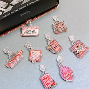 Puede incluir: Una colección de dijes de libros acrílicos con varias frases y gráficos, incluyendo "Hello I'm Never Not Reading Smut", "Smutty Book Club" y "Explicit Content". Los dijes son rosas, rojos y blancos, y están unidos a un lector Kindle.