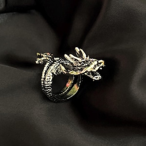 May include: A silver dragon ring with detailed craftsmanship. The dragon design wraps around the finger, showcasing scales, claws, and a detailed head. The ring is set against a dark, textured backdrop, emphasizing the metallic finish and design.