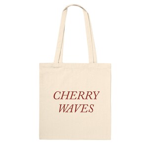 Może przedstawiać: Kremowa torba typu tote z napisem "CHERRY WAVES" wydrukowanym ciemnoczerwoną, stylizowaną czcionką. Torba ma dwa pasujące kremowe uchwyty.