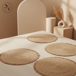 Peut inclure: Quatre dessous de plat ronds en jute de couleur naturelle avec un bord effiloché sont disposés sur une table en bois de couleur claire. Les dessous de plat ont un motif de cercles concentriques. Le fond comprend un vase avec des fleurs séchées et un logo "NOLYAA".