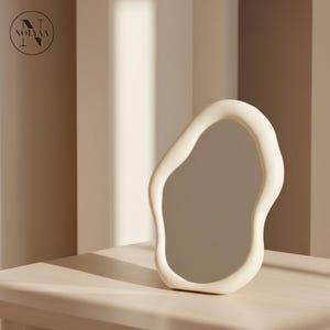 May include: An off-white, organically shaped mirror with a smooth, matte finish. The mirror has a unique, irregular outline and is set against a neutral-toned background. The mirror is placed on a light-coloured surface. The logo "NOLYAA" is in the top left corner.