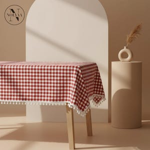 Peut inclure: Une nappe à carreaux rouges et blancs avec une bordure à pompons blancs, drapée sur une table en bois. Un petit vase crème avec de l'herbe séchée repose sur un piédestal cylindrique. L'arrière-plan est beige neutre.