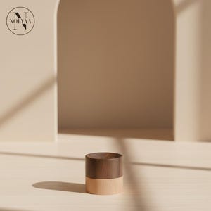 Peut inclure: Un petit récipient cylindrique en bois avec un dessus marron foncé et une base marron clair. Le récipient est posé sur une surface en bois de couleur claire. L'arrière-plan présente une arche aux tons neutres et un logo dans le coin supérieur gauche.