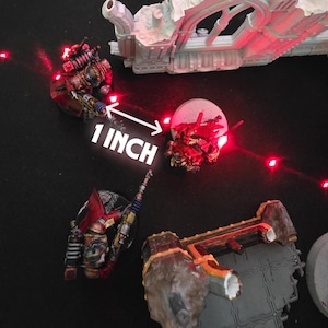 Peut inclure: Figurines miniatures de soldats futuristes, peintes en rouge, or et noir, avec des armures et des armes détaillées. Le texte "1 INCH" est affiché. La scène est éclairée par des lumières rouges, suggérant un thème de science-fiction ou de fantaisie. 2.54 cm.