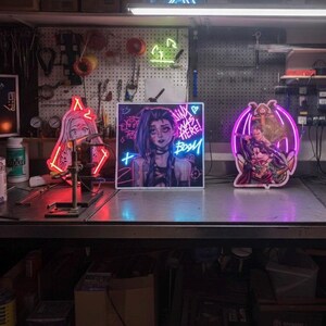 Puede incluir: Una escena de taller con varios letreros de neón. Los letreros incluyen un personaje de estilo anime rojo, un corazón verde, un gesto con la mano azul y un personaje morado con el texto "Jinx was here! Boom". Los letreros están sobre un banco de trabajo de metal.