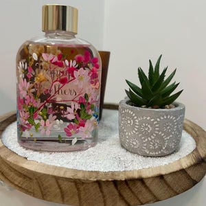 May include: A clear glass bottle with a gold cap, filled with liquid and floral decorations, sits on a wooden tray with white sand. A small potted succulent is next to the bottle. The bottle has the word "Mothers" on it.