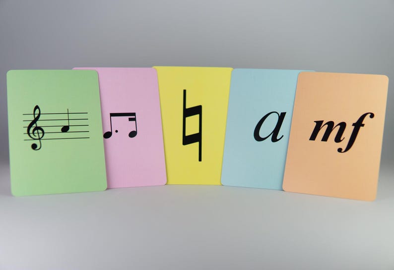 K&ouml;nnte beinhalten: Eine Sammlung farbiger Karteikarten mit musikalischen Symbolen und Begriffen. Die Karten sind in Gr&uuml;n, Rosa, Gelb, Hellblau und Pfirsich gehalten. Jede Karte zeigt ein anderes musikalisches Symbol oder einen Begriff in Schwarz, darunter ein Violinschl&uuml;ssel, eine Viertelnote und die Buchstaben &bdquo;a&ldquo; und &bdquo;mf&ldquo;.