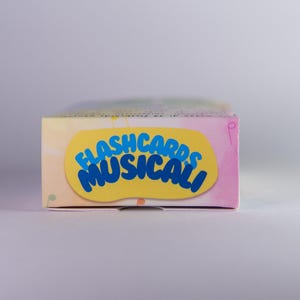 K&ouml;nnte beinhalten: Eine rechteckige Schachtel mit Lernkarten mit dem Text "FLASHCARDS MUSICALI" in Blau auf einem gelben Oval. Die Schachtel hat ein Pastellfarbschema mit rosa, gelben und blauen Farbt&ouml;nen. Der Hintergrund ist eine weiche, neutrale Farbe.