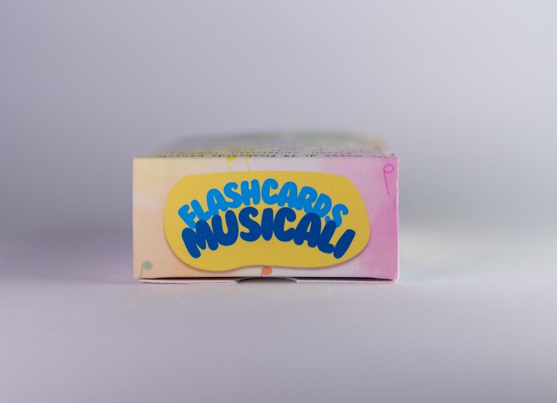 K&ouml;nnte beinhalten: Eine rechteckige Schachtel mit Lernkarten mit dem Text "FLASHCARDS MUSICALI" in Blau auf einem gelben Oval. Die Schachtel hat ein Pastellfarbschema mit rosa, gelben und blauen Farbt&ouml;nen. Der Hintergrund ist eine weiche, neutrale Farbe.