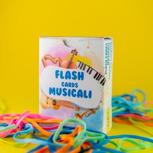K&ouml;nnte beinhalten: Eine Schachtel mit musikalischen Lernkarten mit einem Aquarelldesign mit Harfe, Geige, Klaviertasten, Gitarre und Trompete. Auf der Schachtel steht in blauer Schrift "FLASH CARDS MUSICALI". Bunte Gummib&auml;nder liegen vor einem gelben Hintergrund.