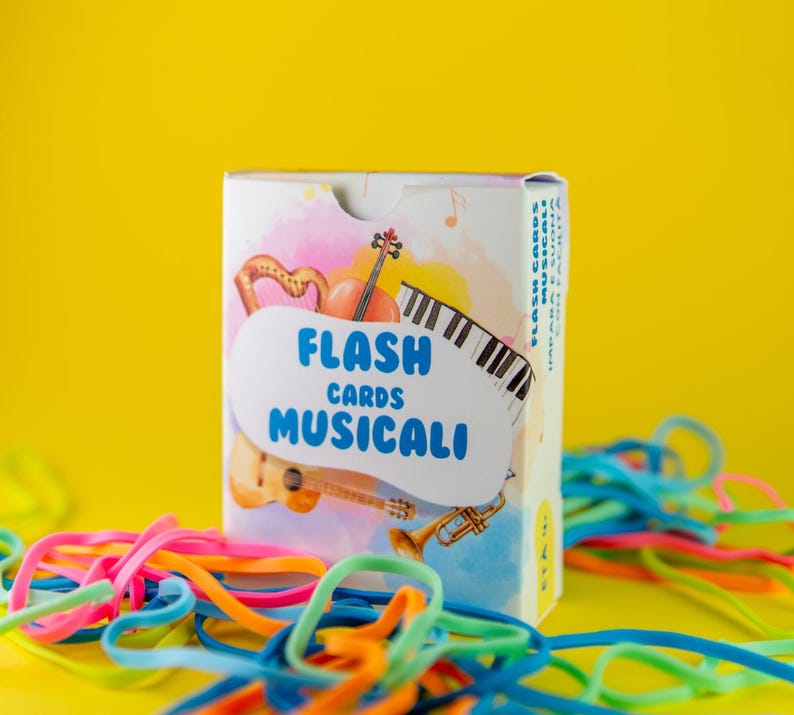 K&ouml;nnte beinhalten: Eine Schachtel mit musikalischen Lernkarten mit einem Aquarelldesign mit Harfe, Geige, Klaviertasten, Gitarre und Trompete. Auf der Schachtel steht in blauer Schrift "FLASH CARDS MUSICALI". Bunte Gummib&auml;nder liegen vor einem gelben Hintergrund.
