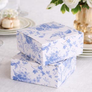 May include: Two stacked white gift boxes with a blue floral pattern. The boxes are on a white tablecloth, with wine glasses and a floral arrangement in the background. The boxes have a classic, elegant design.