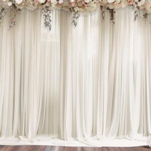May include: A sheer white curtain backdrop with a floral arrangement at the top. The flowers are a mix of white and pink, with greenery accents. The curtain is draped and creates a soft, elegant look, suitable for events.