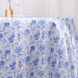 May include: A white tablecloth with a blue floral pattern. The design features detailed blue flowers and foliage, creating a classic and elegant look. The tablecloth is draped over a table, suggesting its use for dining or special events.