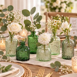 May include: A collection of five small glass vases in varying shades of green, each featuring embossed patterns. The vases are filled with assorted white flowers, including roses, daisies, and eucalyptus sprigs, arranged on a table.