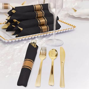 May include: A set of black napkins wrapped in gold bands, accompanied by gold-colored plastic forks, spoons, and knives. The napkins are stacked on a white rectangular tray with a beaded edge, next to a clear glass stemware piece.