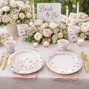 May include: A beautifully arranged table setting for a bridal shower. White plates and cups are adorned with floral designs. Pink napkins, gold cutlery, and candles complement the centerpiece of flowers. A sign reads "Bride to Be".