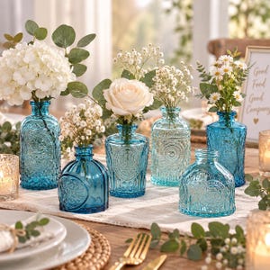 May include: A collection of six blue glass vases with embossed patterns, each holding various white flowers including hydrangeas, roses, and baby's breath. The vases vary in shape and size, featuring intricate circular and floral designs.