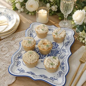 May include: Six cupcakes with beige frosting and green accents are arranged on a white and blue patterned serving tray. The tray has a scalloped edge. A lit candle, a wine glass, and flowers are in the background. Some cupcakes have a small tag that says "especially GIFT".