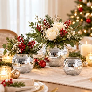 May include: Two spherical disco ball vases filled with white roses, white chrysanthemums, green pine branches, eucalyptus leaves, and red berries. A smaller empty spherical disco ball vase sits nearby, surrounded by candles and holiday decor.