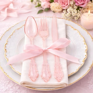 May include: A place setting with pink plastic cutlery, including a spoon, fork, and knife, tied with a pink ribbon on a white napkin. The plate has a gold rim. A lit candle and flowers are in the background.