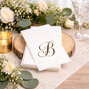 May include: A stack of white paper napkins with a gold letter "B" printed on the top napkin. The napkins are on a wooden slice, with a lit gold candle and floral arrangements in the background.