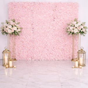May include: A large square backdrop covered in dense light pink artificial flowers. Two tall gold metal stands hold floral arrangements of white and pink roses and greenery. Four gold-framed lanterns with lit white pillar candles sit below.