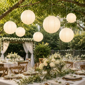 May include: A collection of round, off-white paper lanterns hanging from tree branches above an outdoor dining area. The lanterns feature a ribbed texture and emit a soft glow, suspended at varying heights over tables set with white linens.
