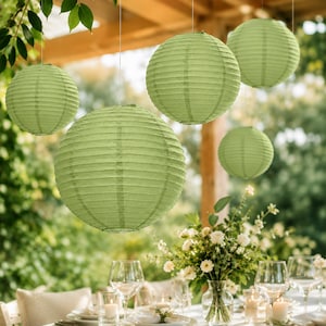 May include: Five spherical paper lanterns in a soft sage green color are suspended at varying heights. Each lantern features a ribbed texture created by thin wire framing beneath the paper surface, providing a rounded shape for outdoor lighting.