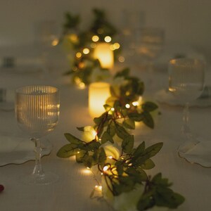 May include: A table setting with two clear wine glasses, a garland of white flowers and green leaves, and small string lights. Two glowing pillar candles are in the background, creating a warm, inviting ambiance.